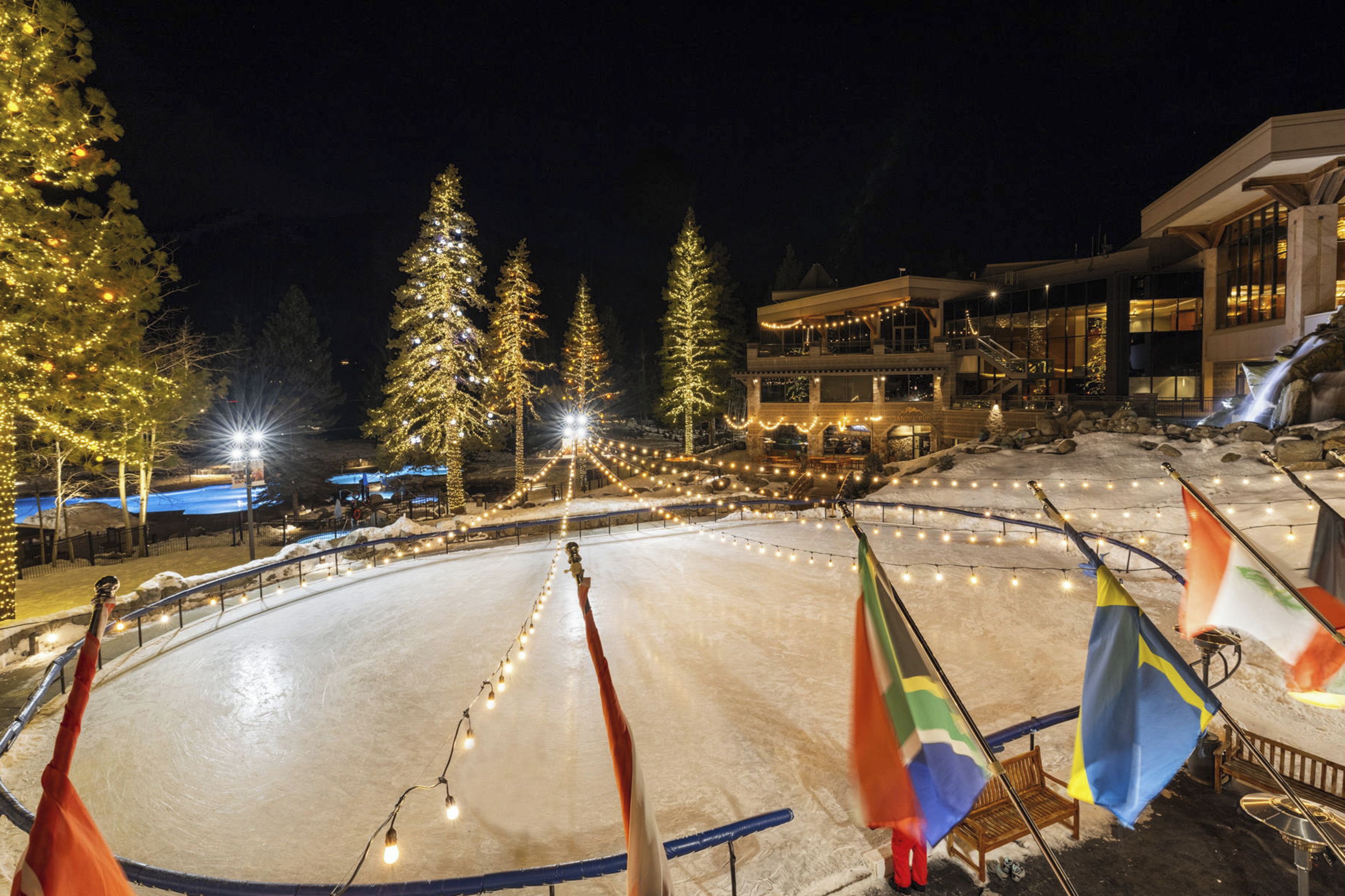 An ice rink with flags, trees, and a building in the background.