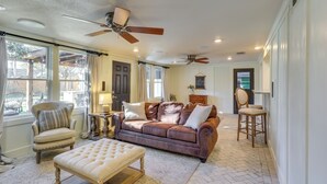 A spacious living room with a brown leather sofa, a tufted ottoman, a wooden chair, and a ceiling fan. - 1 Mi to Texas Tech: Group Getaway w/ Pool & Sauna! (Lubbock)
