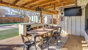 An outdoor patio with a wooden table and chairs, a TV mounted on a wall, and a wooden ceiling with hanging lights. - 1 Mi to Texas Tech: Group Getaway w/ Pool & Sauna! (Lubbock)