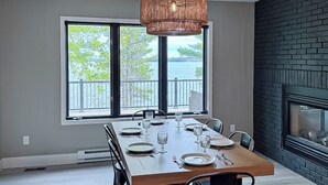 A dining area with a large table set for six, a fireplace, and a view of a lake through large windows. - Luxury Rainy Lake House With Private Dock! (International Falls)