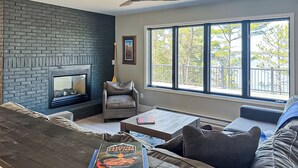 A living room with a fireplace, a large window with a view, a sofa, and a coffee table. - Luxury Rainy Lake House With Private Dock! (International Falls)