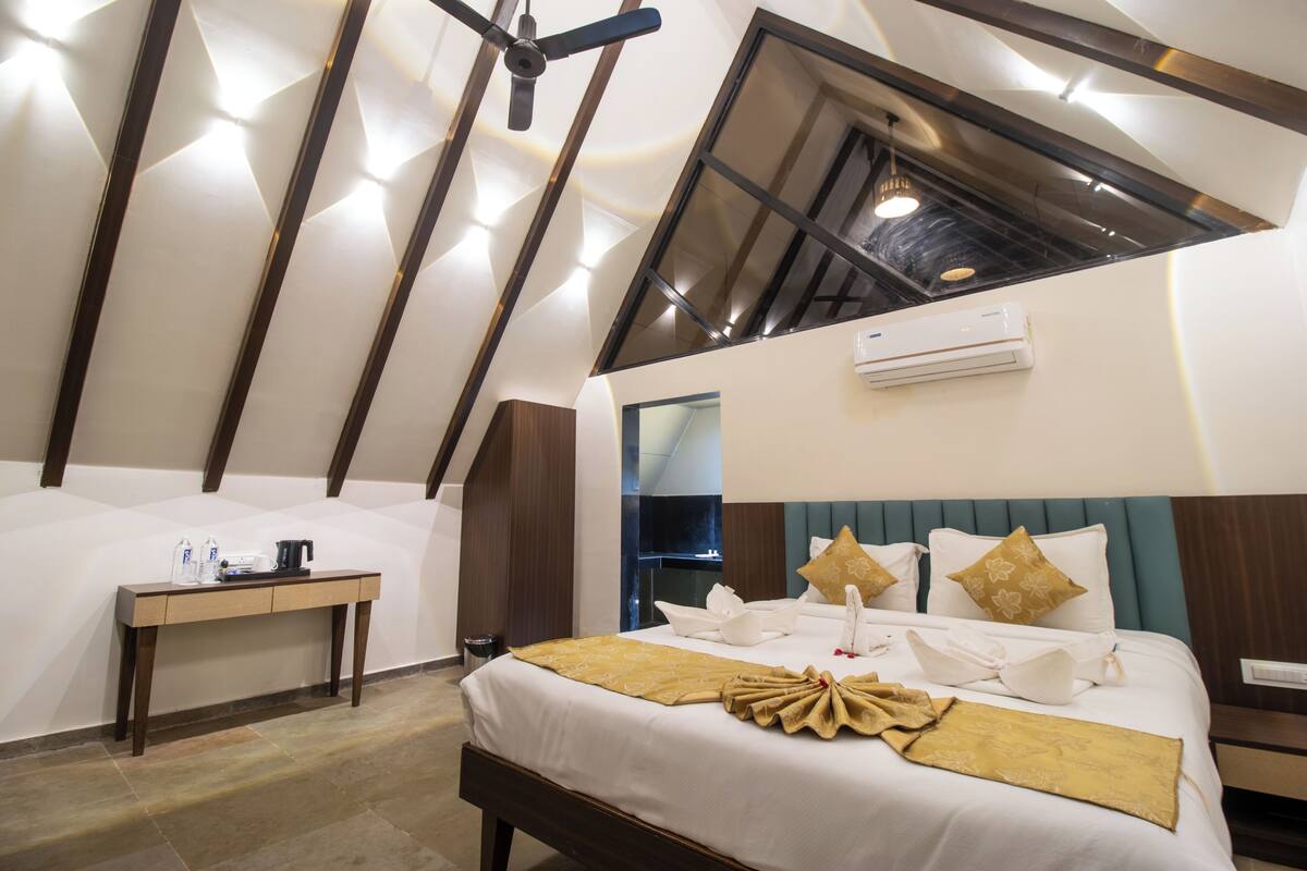 Ein Zimmer mit hohen Decken und einem Oberlicht, ein Bett mit weißen Leinen und goldenen Akzenten, ein Nachttisch mit Wasserkocher und ein kleiner Schreibtisch mit Stuhl.