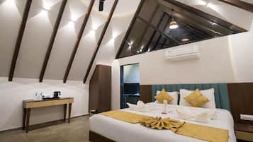 Habitación de hotel moderna con techo inclinado, cama con cubrecama dorado, mesita de noche con tetera y unidad de aire acondicionado.