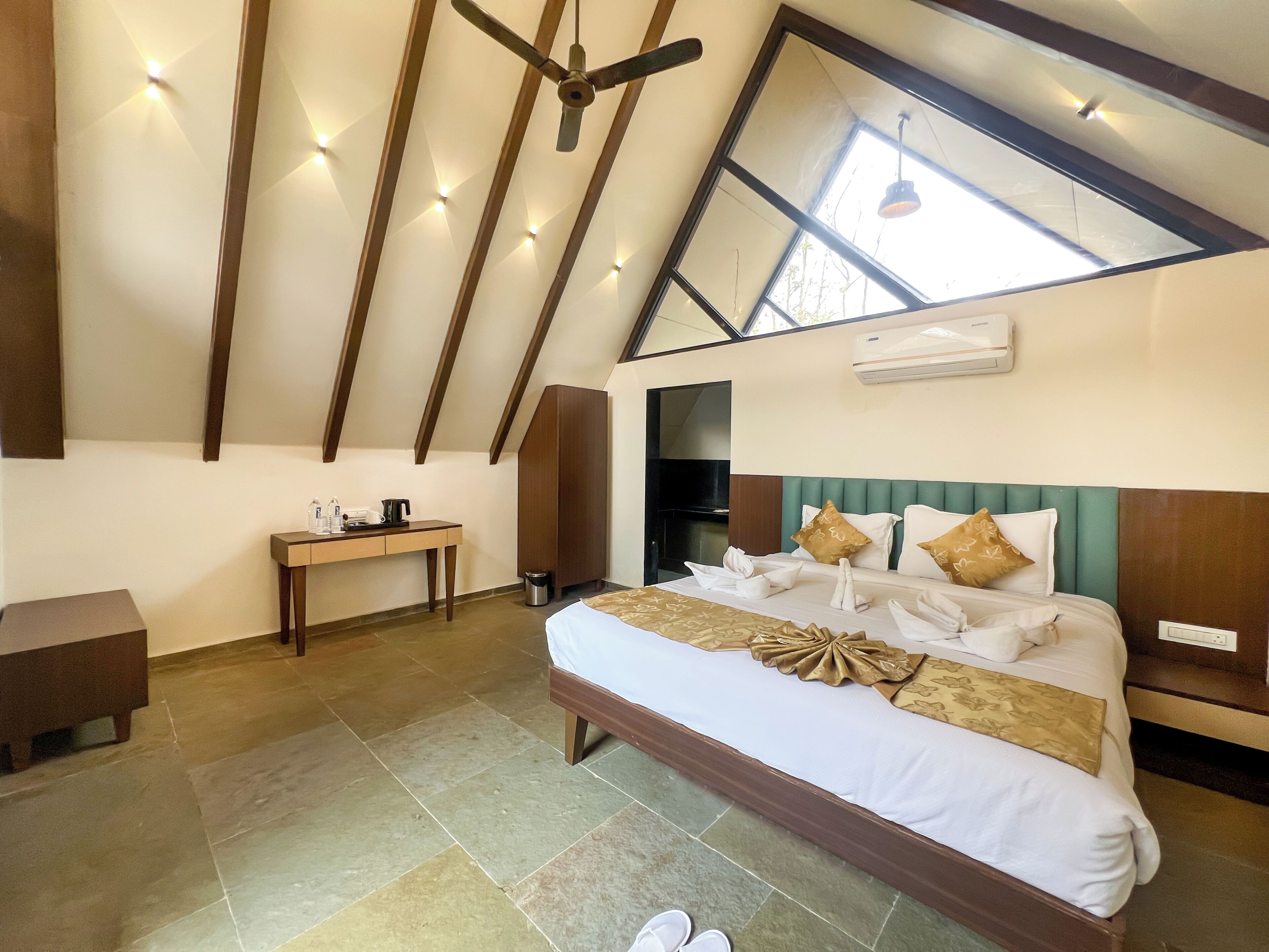 A hotel room with a high ceiling, a skylight, a bed with neatly arranged towels, a bedside table, and a small cabinet.