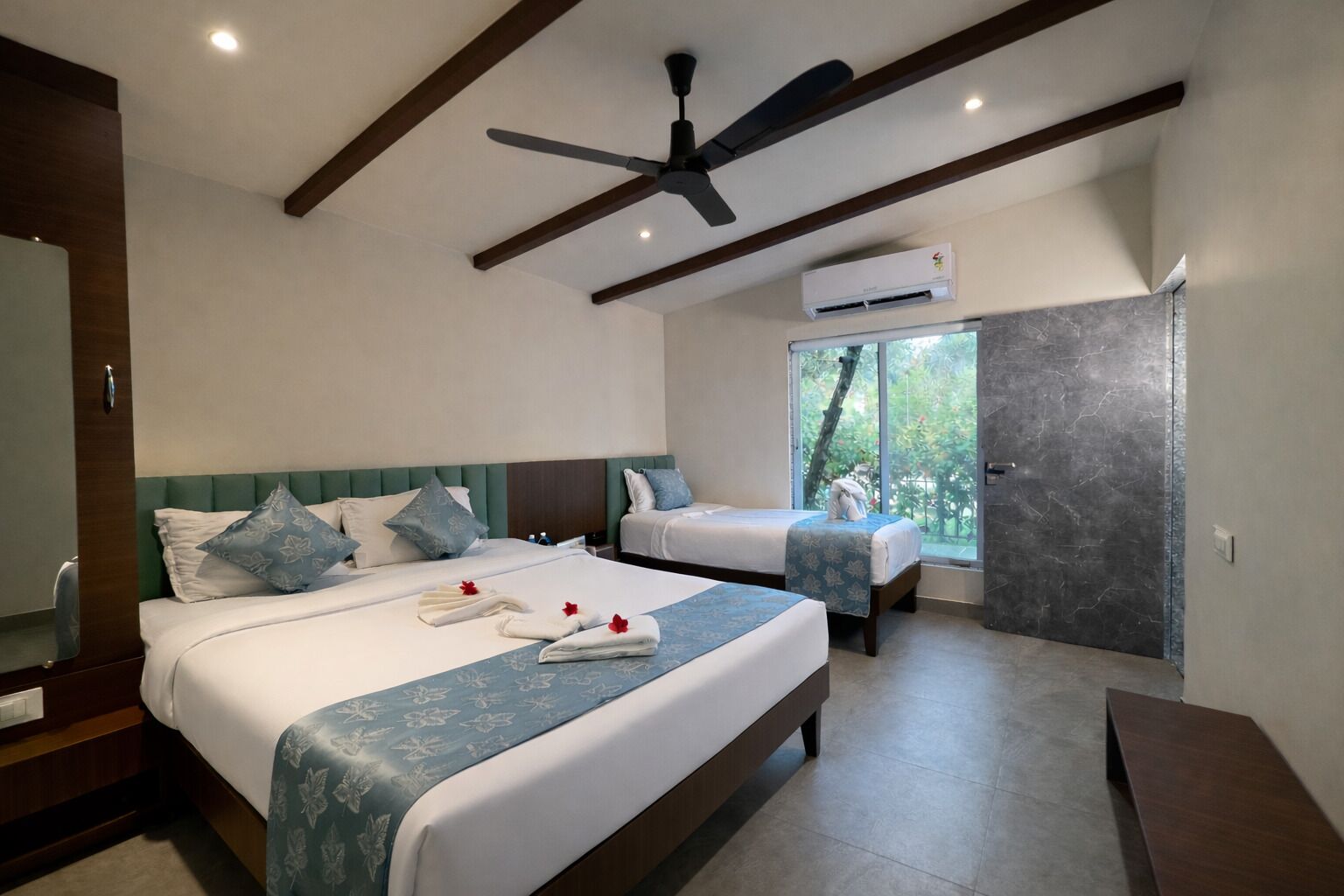 Quarto de hotel com duas camas, ventilador de teto, ar condicionado e vista para vegetação pela janela.