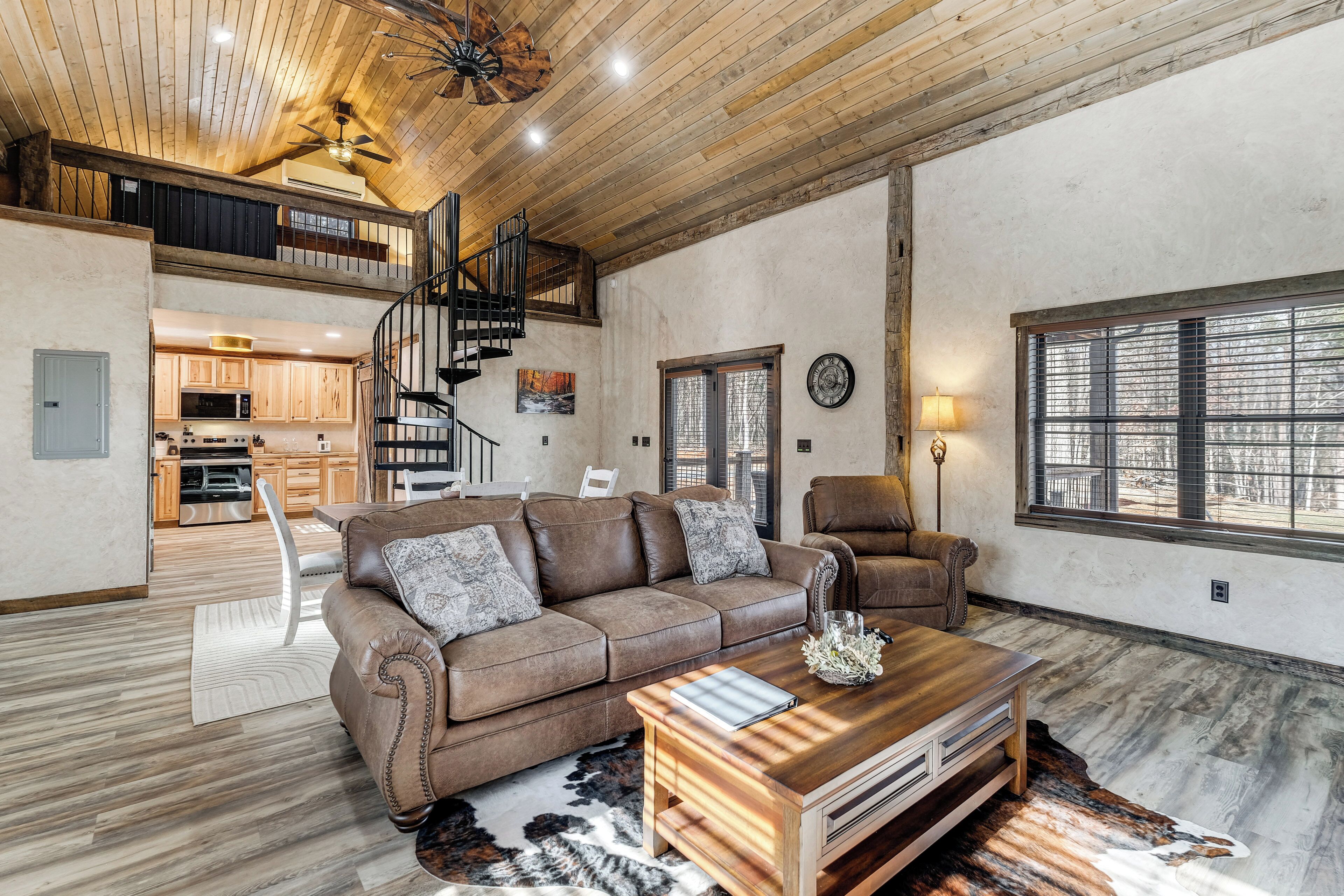 A spacious living area with a brown leather sofa, a wooden coffee table, and a large window. A staircase leads to an upper level with a kitchen.