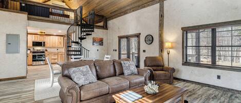 A spacious living area with a brown leather sofa, a wooden coffee table, and a large window. A staircase leads to an upper level with a kitchen.