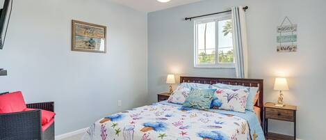 Un dormitorio con una cama, dos mesitas de noche, una silla, un ventilador de techo y una ventana con cortinas.