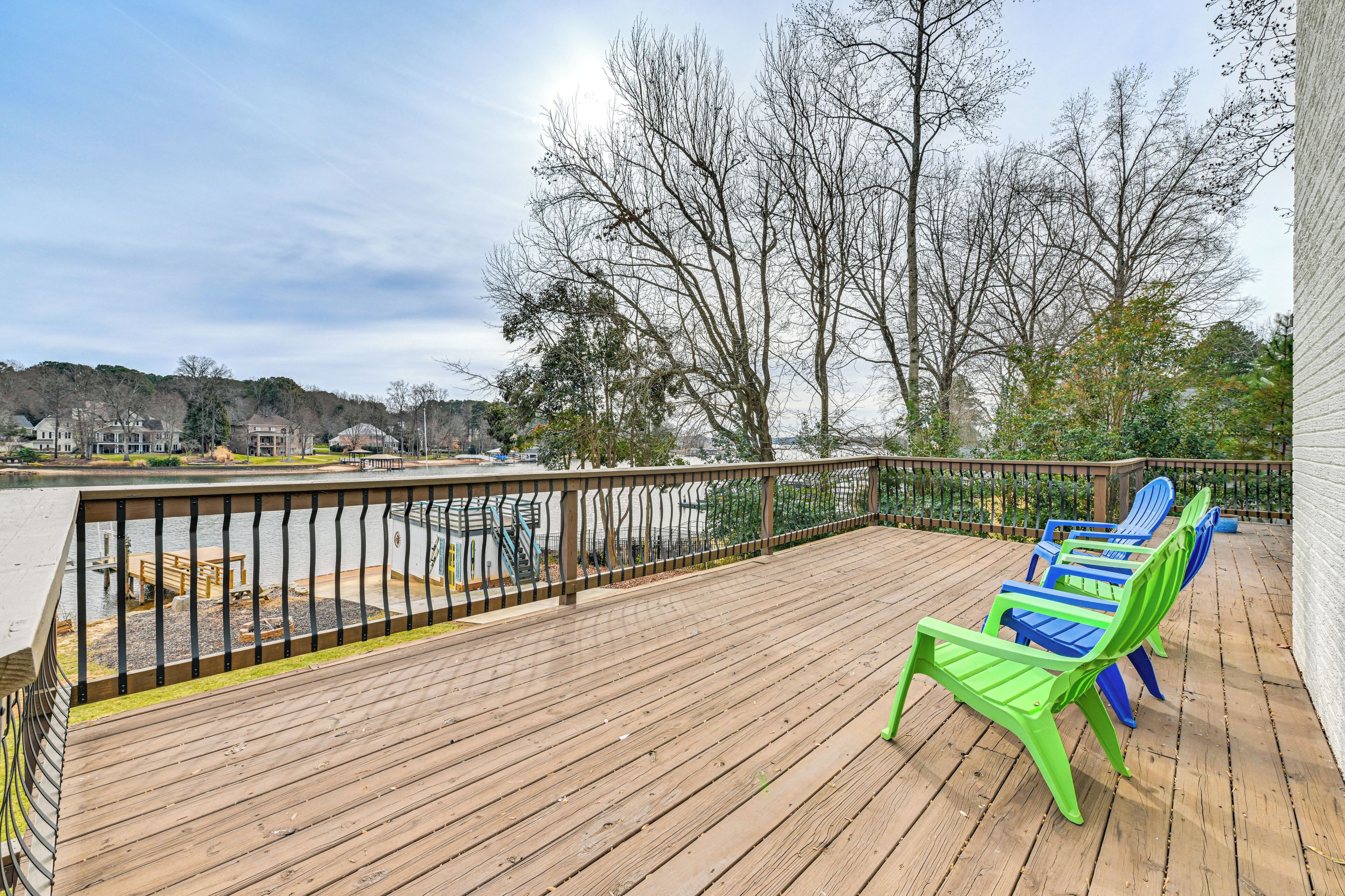 A wooden deck with a railing, green and blue Adirondack chairs, and a view of a parking lot and trees.