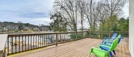 A wooden deck with a railing, green and blue Adirondack chairs, and a view of a parking lot and trees.
