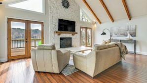 A spacious living room with a stone fireplace, a television mounted on the wall, and a balcony with a view of a lake. - Boat Dock & Game Room: Waterfront Lake Norman Gem (Mooresville)