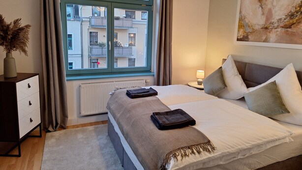 Un dormitorio moderno con una cama grande, una mesita de noche con lámpara, un radiador y vistas a un edificio residencial a través de una ventana.