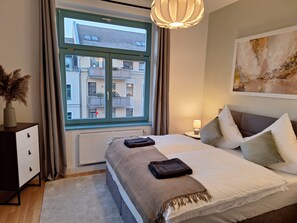 A modern bedroom with a large bed, a bedside table with a lamp, a radiator, and a view of a residential building through a window. - Stylish accommodations in the historic district (Chemnitz)