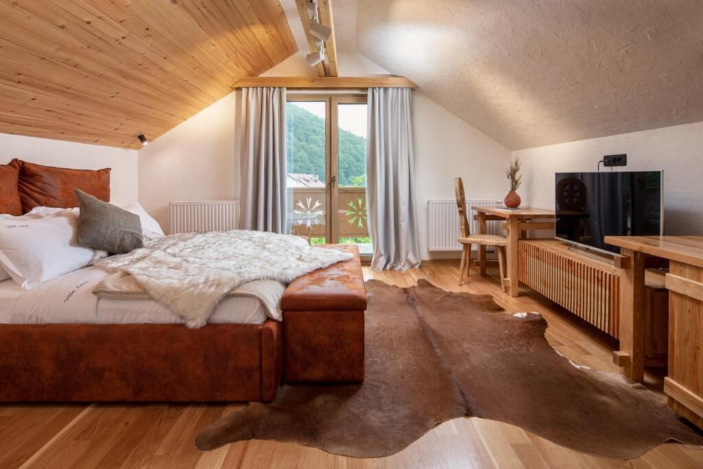 A bedroom with a wooden ceiling, a bed with a white blanket, a wooden table, a television, and a view of the outdoors.