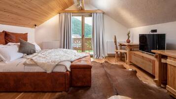 A bedroom with a wooden ceiling, a bed with a white blanket, a wooden table, a television, and a view of the outdoors.