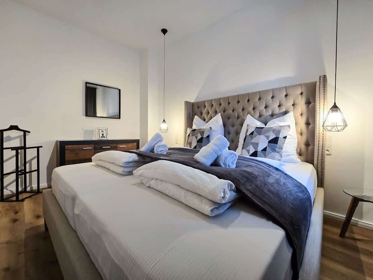 A neatly made bed with pillows and a blanket, a bedside table with a lamp, and a small table with a chair in a modern bedroom.