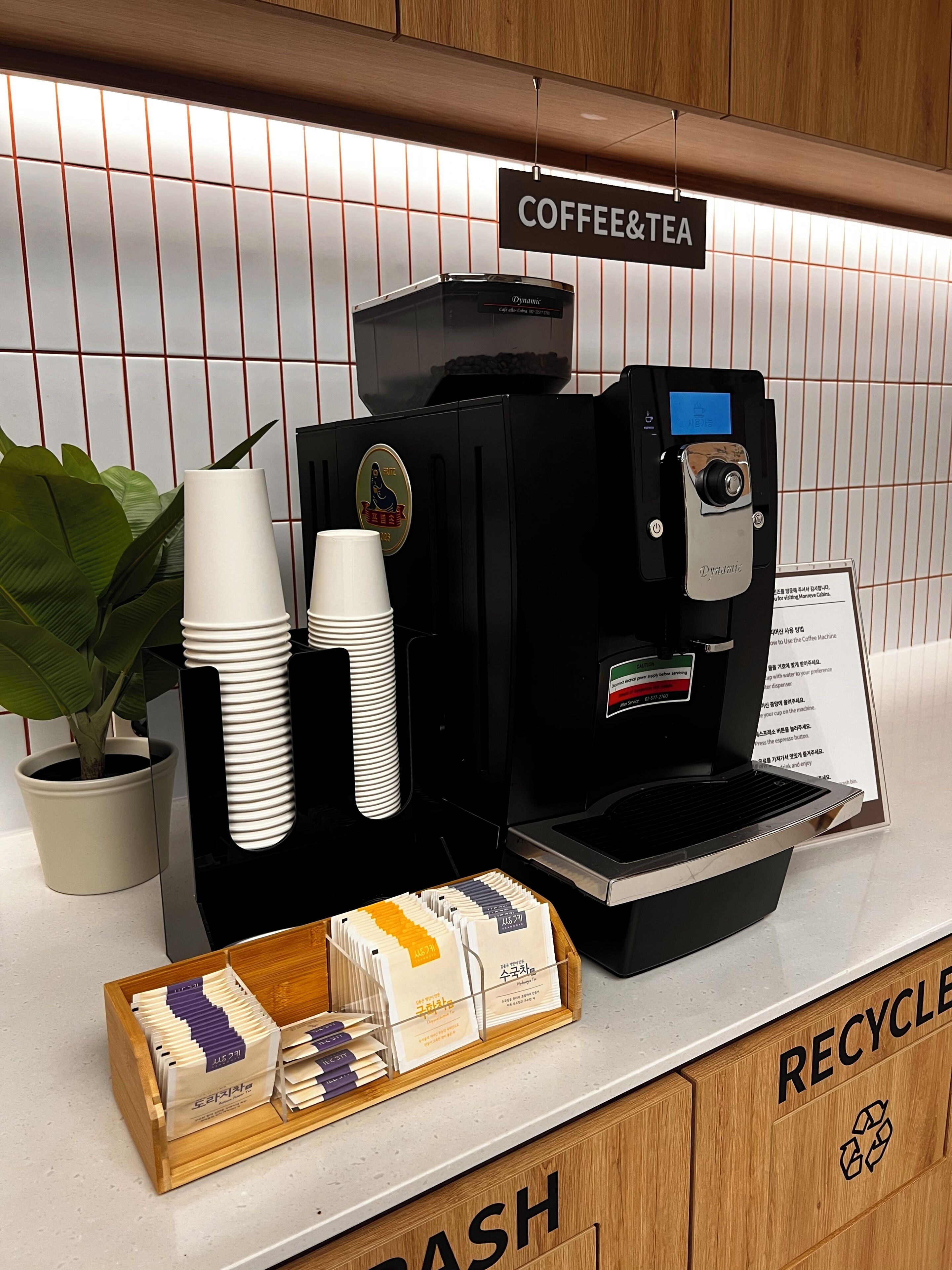 A coffee and tea station with a coffee machine, disposable cups, and a recycling bin.