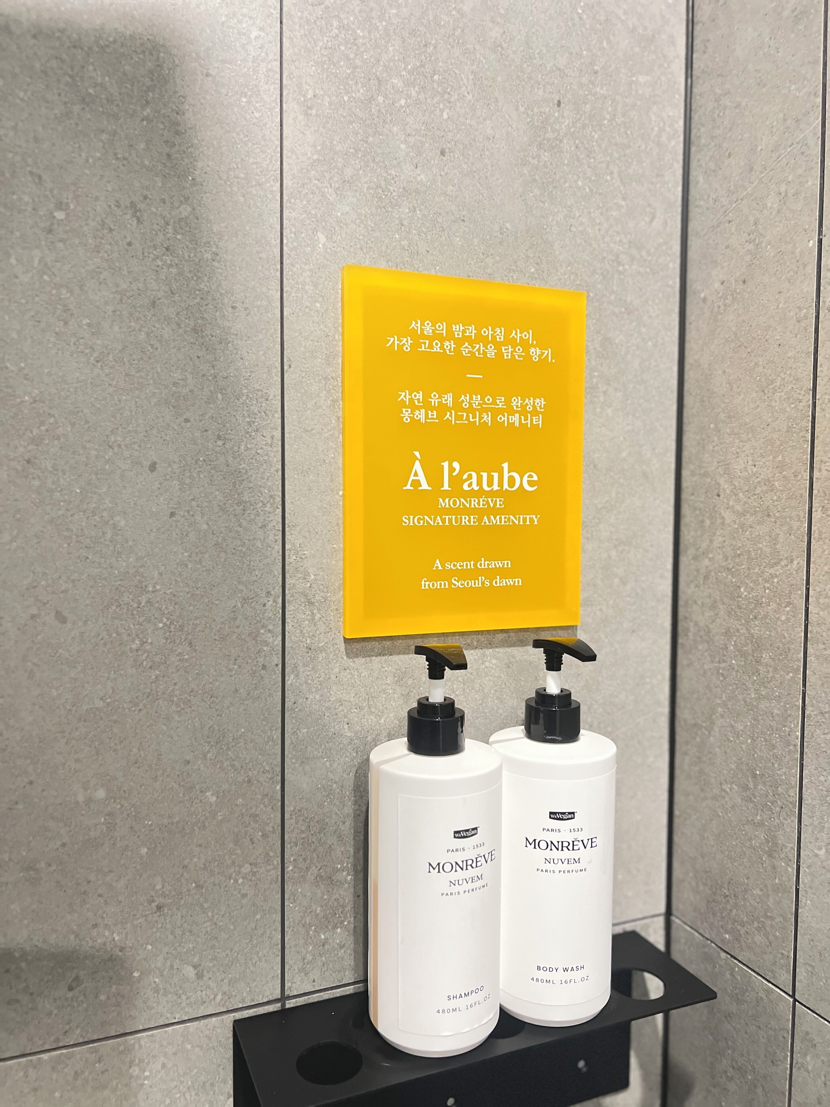 Un baño con un letrero amarillo, dos botellas de champú Monréve y un dispensador de jabón negro.