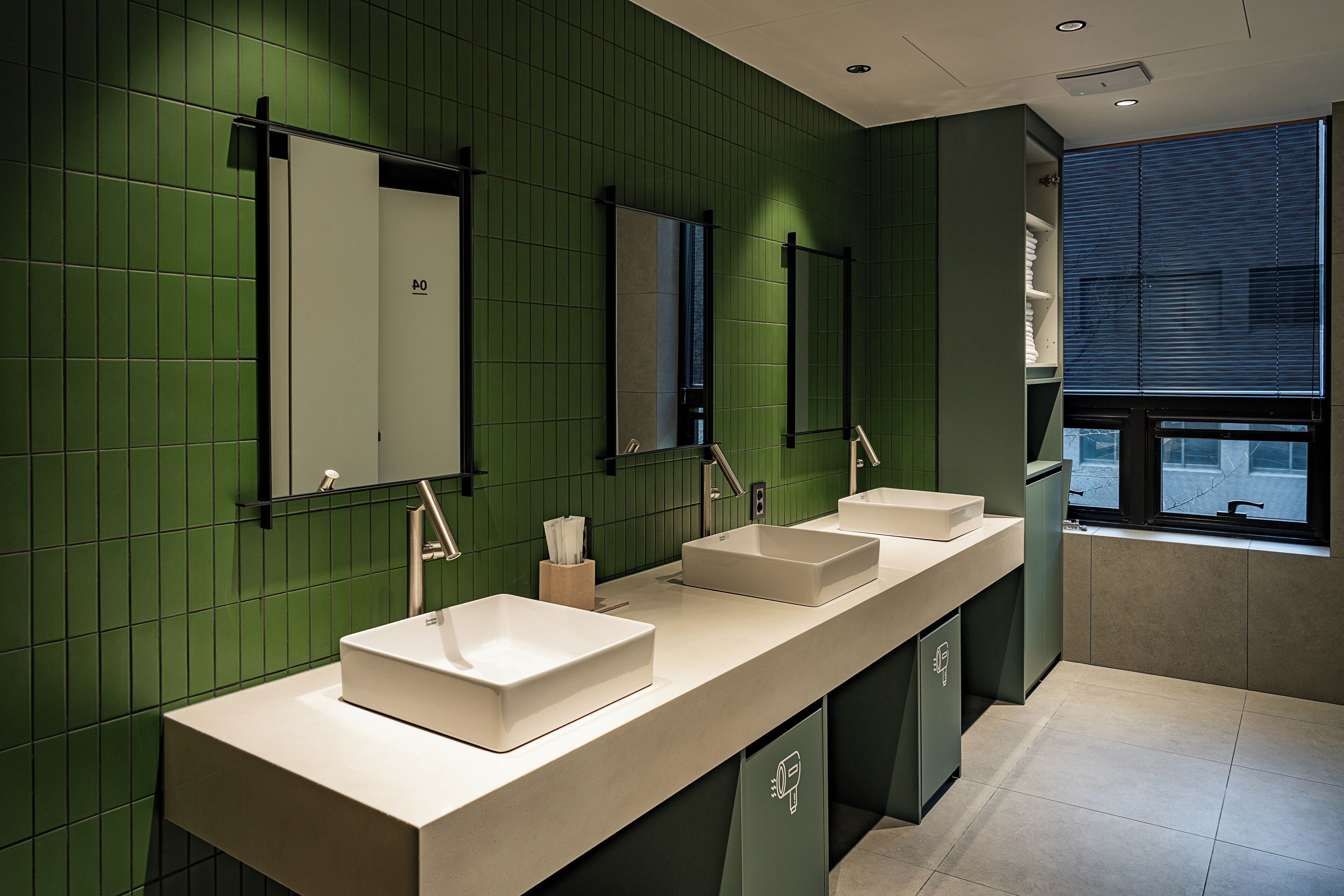 Un baño moderno con tres lavamanos, paredes de azulejos verdes y una ventana con persianas.