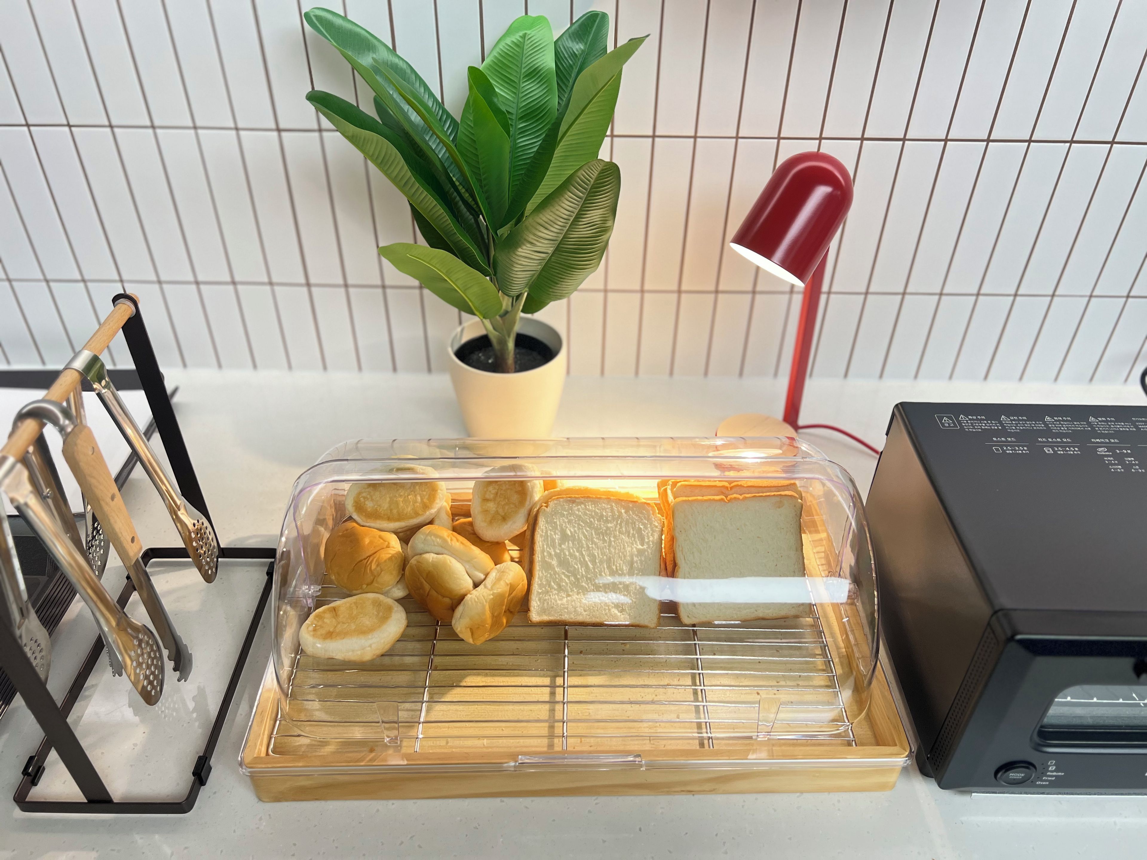 A kitchen counter with a bread box, a plant, a red lamp, and a grater.