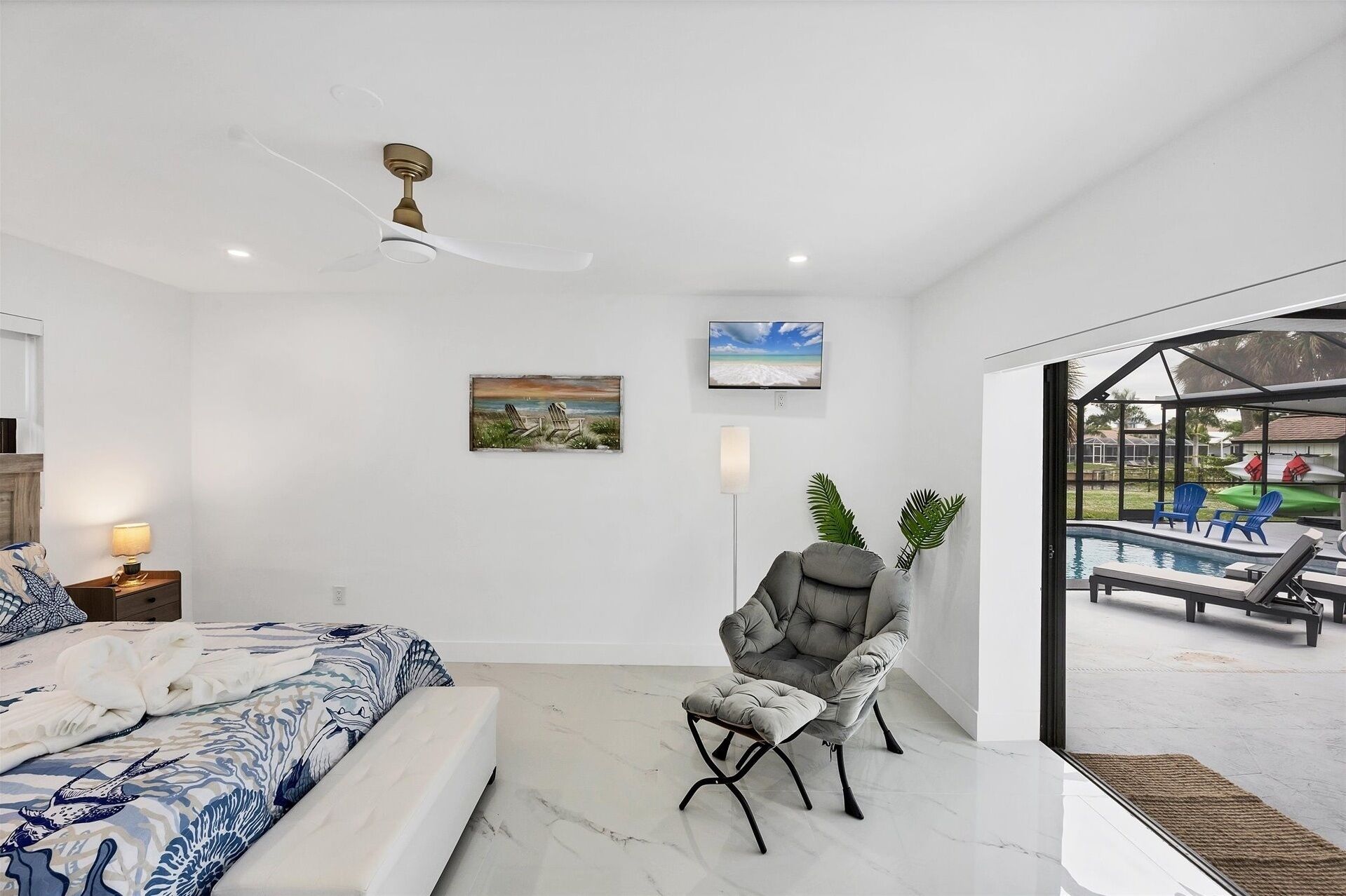 A bedroom with a bed, a chair, a painting, a television, and a view of an outdoor pool area.