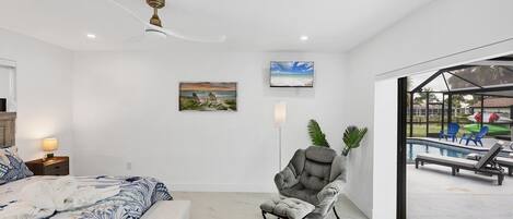 A bedroom with a bed, a chair, a painting, a television, and a view of an outdoor pool area.