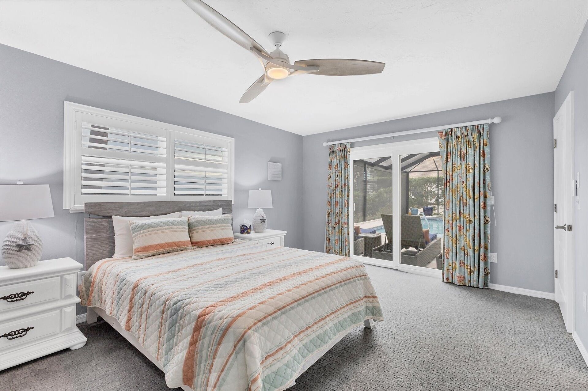 A bedroom with a bed, nightstands, a ceiling fan, a window with blinds, and a view of a pool.