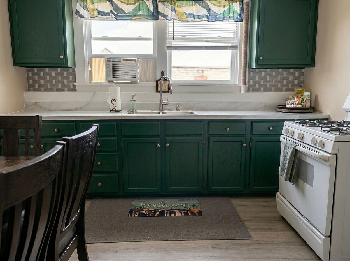 Une cuisine avec des meubles de couleur verte, une cuisinière, un évier et une fenêtre avec des stores.