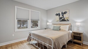 Une chambre à coucher avec un lit, des tables de chevet, une fenêtre avec des stores et un tableau au mur.