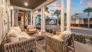 A covered outdoor seating area with sofas and chairs, a small table, and a view of a street with palm trees and buildings. - Beach Bike Cottage 5 Bedroom, King Beds, Coastal Luxury Living, EV Charger (Seacrest Beach)