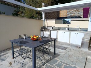 An outdoor kitchen area with a dining table and chairs, a fruit bowl, and a stone-tiled patio. - Bella vista (Santa-Maria-di-Lota)