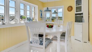 Uma sala de jantar com uma mesa branca, cadeiras e luminárias pendentes. Há janelas com molduras brancas e uma vista para o exterior.