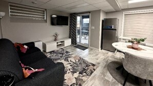 Ein kompakter Wohnraum mit Sofa, kleinem Esstisch, Flachbildfernseher, Kühlschrank und einem Balkon mit Blick.