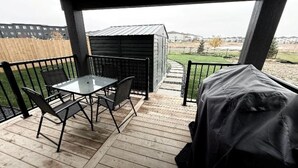 Eine überdachte Terrasse mit Tisch und Stühlen, eine kleine Werkstatt und Blick auf einen eingezäunten Garten sowie Gebäude in der Ferne.