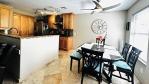 Uma cozinha com armários de madeira, uma ilha central, uma mesa de jantar com cadeiras, um ventilador de teto e um relógio na parede.