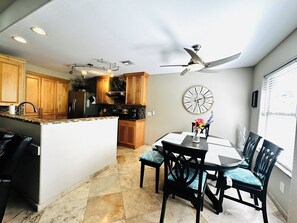 A kitchen with wooden cabinets, a central island, a dining table with chairs, a ceiling fan, and a clock on the wall. - Coastal escape/pool/pickleball/pets/tennis/golf/beach & fun (Laguna Vista)