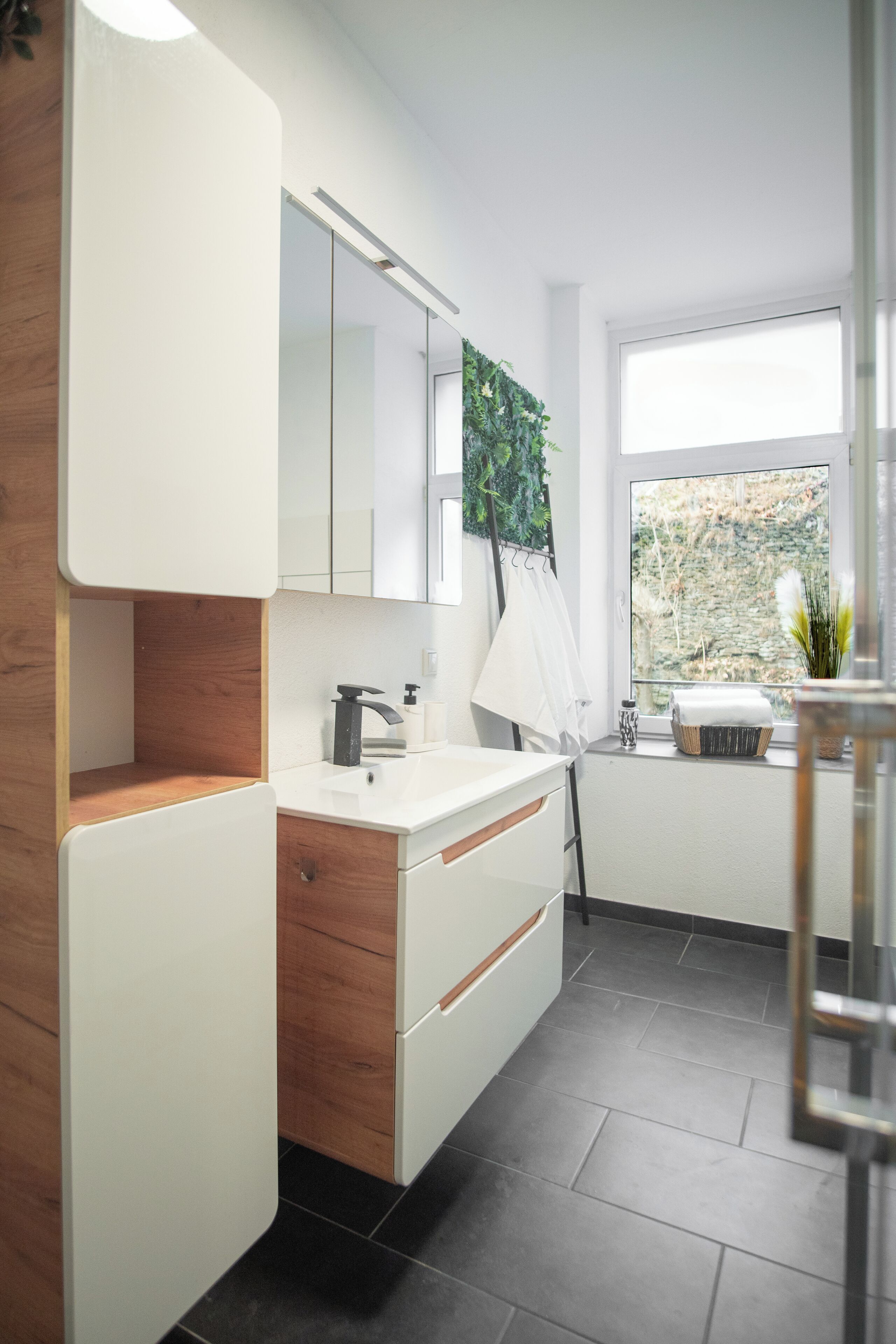 Un baño moderno con tocador blanco, detalle de madera, espejo grande y ventana con vista a árboles.
