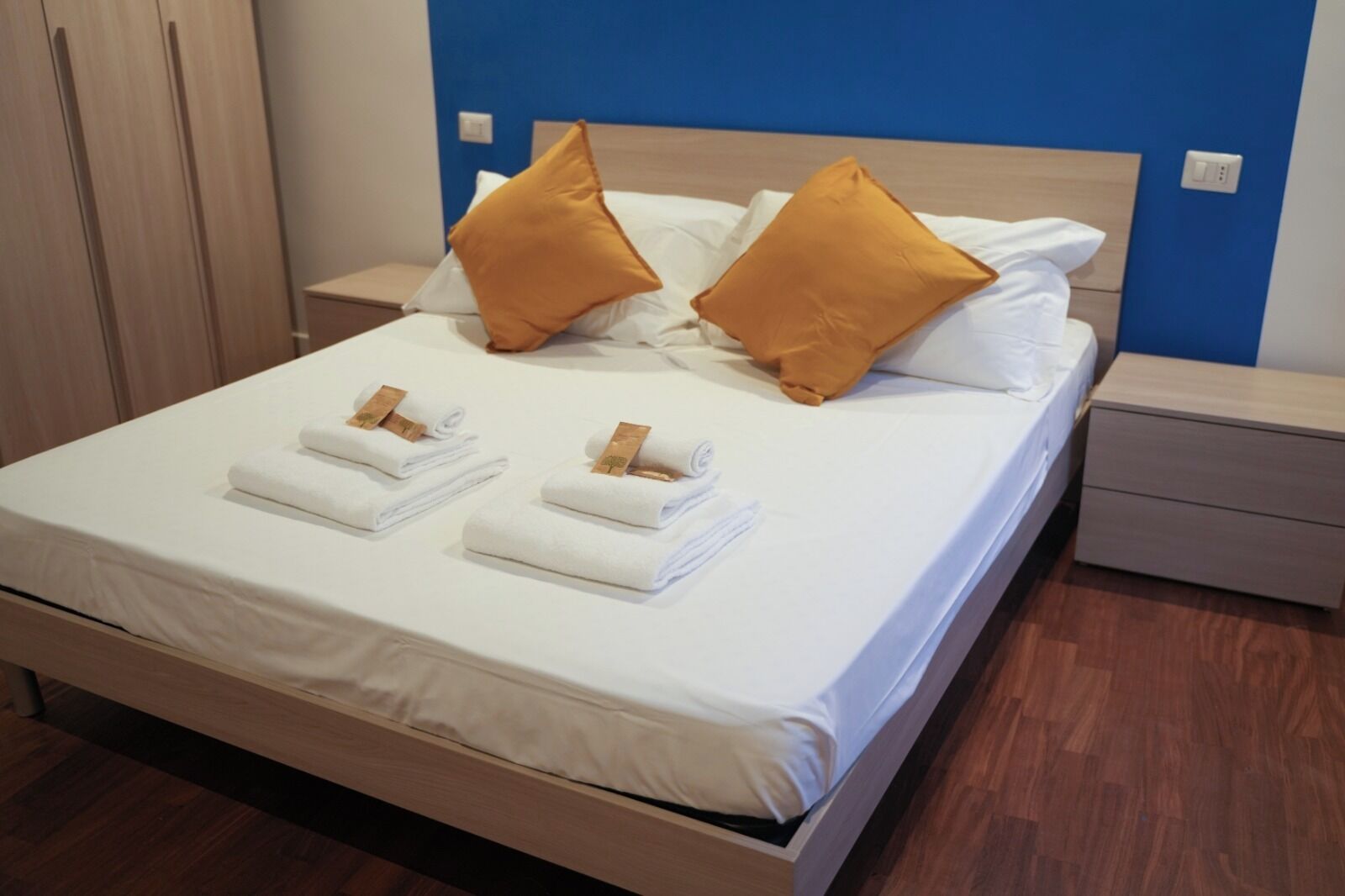 Ein ordentlich bezogenes Bett mit weißen Bettwäsche und zwei braunen Kissen, ein Nachttisch aus Holz mit Schublade und eine blaue Akzentwand.
