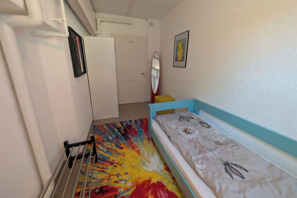 A narrow hallway with a bed on the right, a mirror on the wall, and a colorful rug.