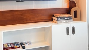 Een moderne woonkamer met een grote flatscreen televisie aan een houten wand, een witte tv-meubel eronder en een decoratieve vaas met gedroogde planten.