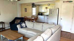 Ein Wohnzimmer mit einem beigefarbenen Ecksofa, einem blauen Sessel und einem Holztisch. Es gibt eine Essecke mit einem Holztisch und Stühlen, sowie eine Küche mit weißen Schränken und Geräten.