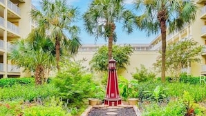 A courtyard with a central red lantern, surrounded by palm trees and potted plants, with a building in the background. - Oceanfront condo - walk to the beach 10min or use free shuttle (Hilton Head Island)