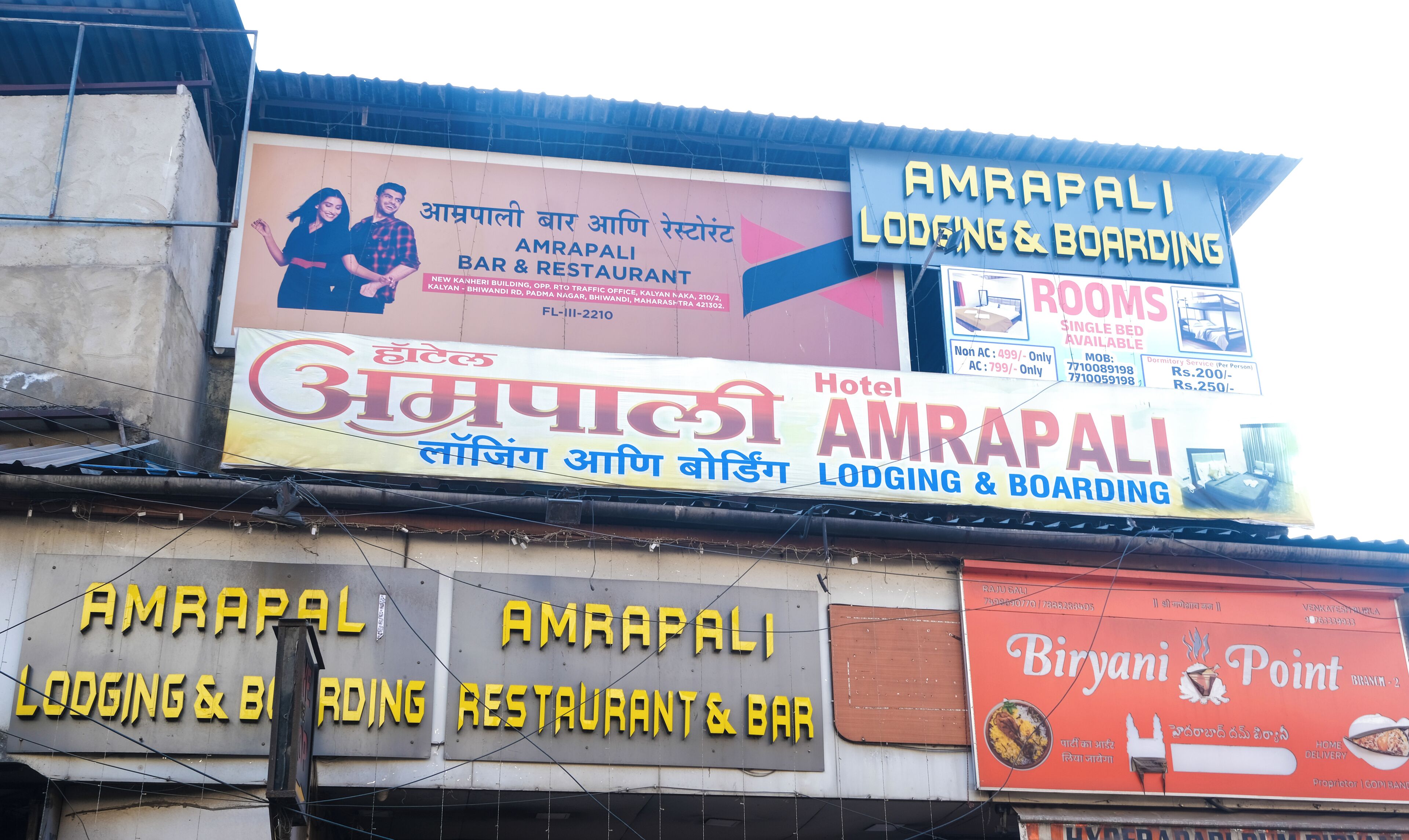 Diversos sinais indicando o alojamento e o restaurante AMRAPALI, incluindo um sinal de hotel e um anúncio do Biriyani Point.