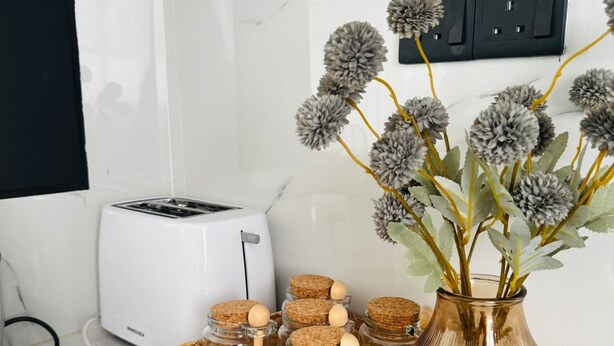 Eine Küchenarbeitsplatte mit einem weißen Toaster, einem gewebten Korb mit Gläsern und einer Vase mit getrockneten Blumen.
