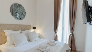 Una cama bien tendida con sábanas blancas y toallas dobladas, un adorno circular en la pared, una lámpara de noche y una ventana con cortinas.