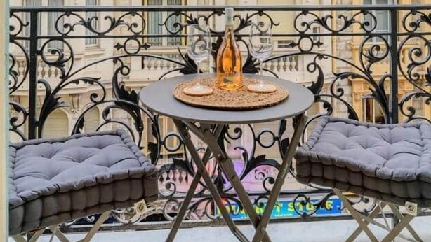Un balcón con una mesa redonda, dos sillas acolchadas y barandillas de hierro forjado decorativo.
