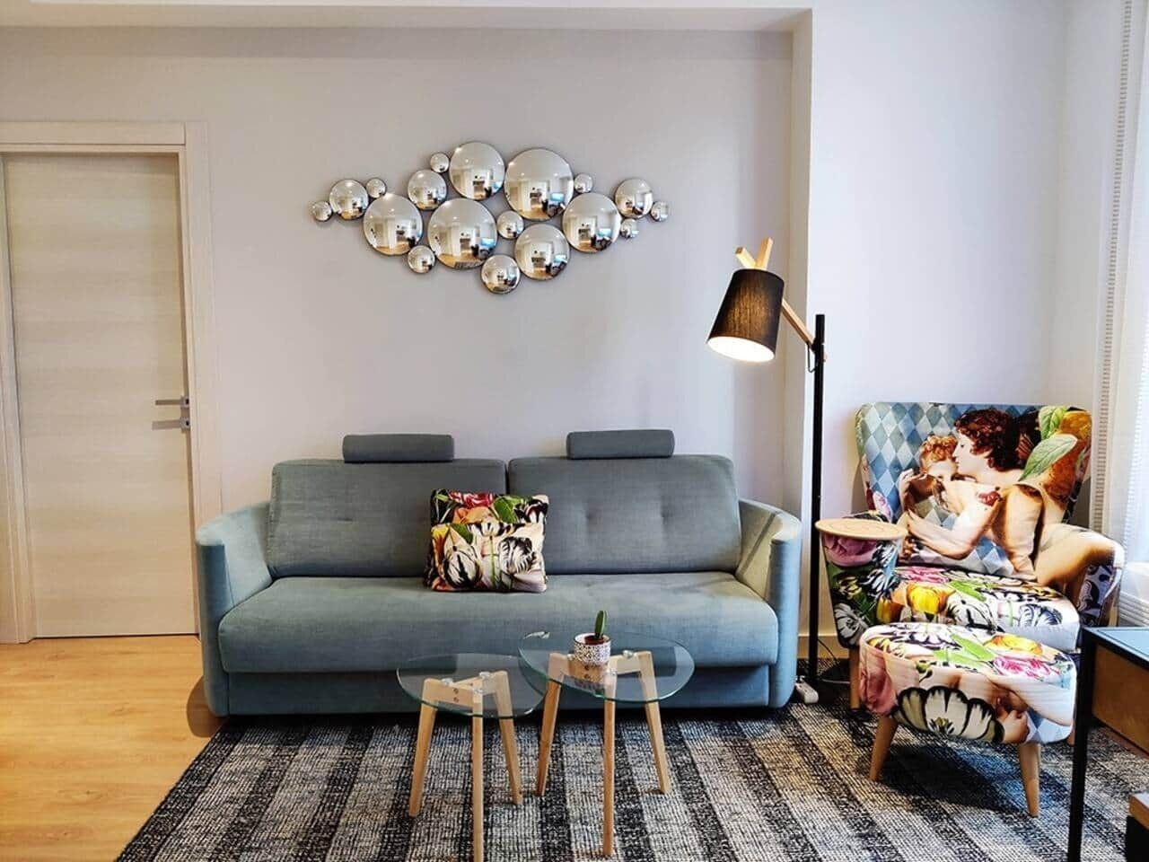 Un salon avec un canapé gris, un fauteuil à motifs, une table basse en verre et une œuvre d’art murale.