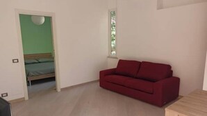 A room with a red sofa, a wooden table, a bed with green bedding, and a window with a view of greenery. - Cozy ground-floor mini apartment a few meters from Rimini's dockyard (Rimini)