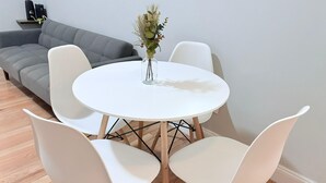 Une table ronde blanche, entourée de quatre chaises blanches, un vase contenant des plantes séchées et, au fond, un canapé gris.