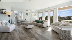 A spacious living area with a wooden floor, a white sofa, a beige sofa, and a dining table with chairs. There is a large window with a view of palm trees and a balcony. - Crow's Nest at Blue Mountain Beach - Ocean View - Private Beach Access (Santa Rosa Beach)